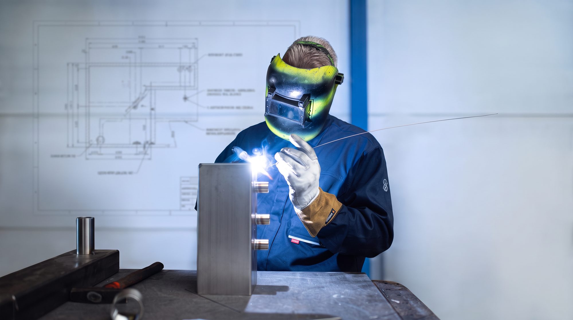 TIG-lasser in blauwe overall met groen lasmasker werkt aan een roestvaststalen werkstuk